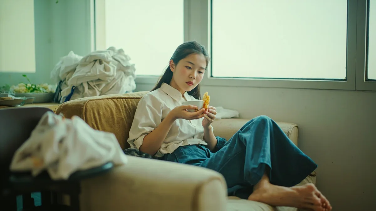 A young adult reclines on a sofa, reaching for a snack instead of doing chores, illustrating “fond of eating and lazy” in Chinese: 好吃懒做 (hào chī lǎn zuò)