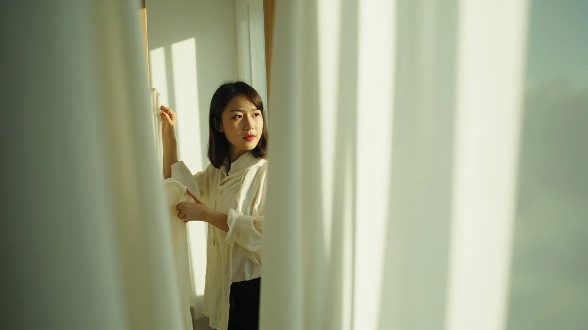 A shopper gestures while trying on a shirt in a fitting room, illustrating “do you have a bigger size” in Chinese: 有大一号的吗？ (yǒu dà yī hào de ma?)