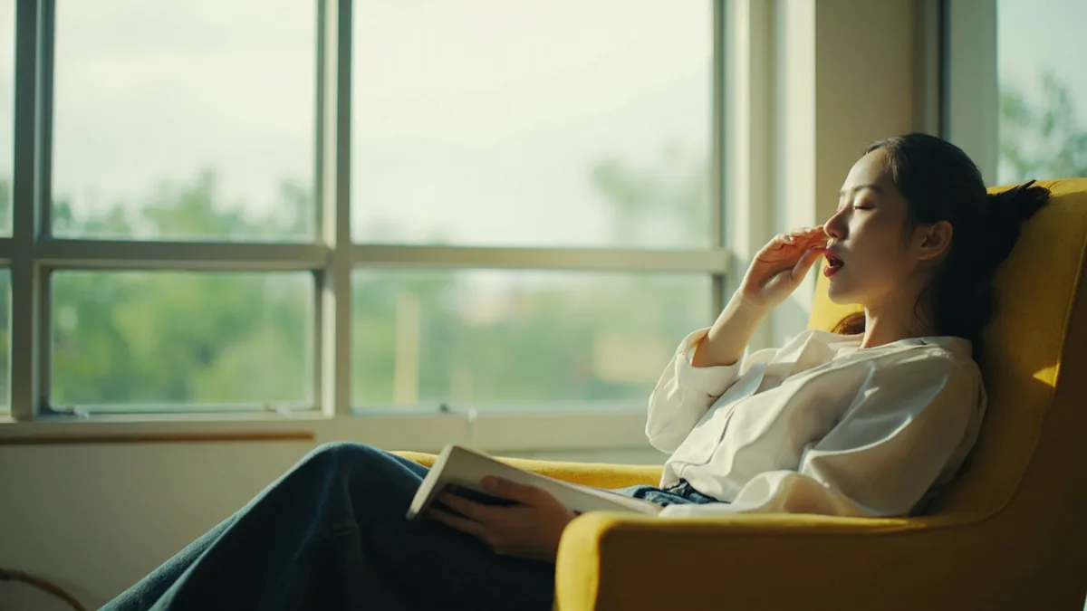 A person yawns while reading a book in a comfortable armchair, illustrating “I'm sleepy” in Chinese: 我困了 (wǒ kùnle)