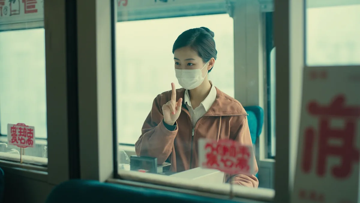 A traveler holds up one finger while speaking to a transit agent, illustrating “one ticket, please” in Chinese: 我要一张票 (wǒ yào yì zhāng piào)