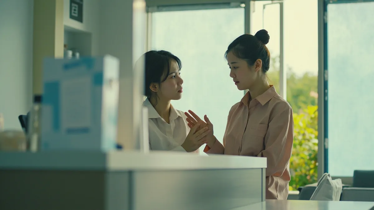 An adult feeling unwell gestures to their stomach while speaking to a clinic receptionist, illustrating “please call a doctor” in Chinese: 请找医生 (qǐng zhǎo yīshēng)