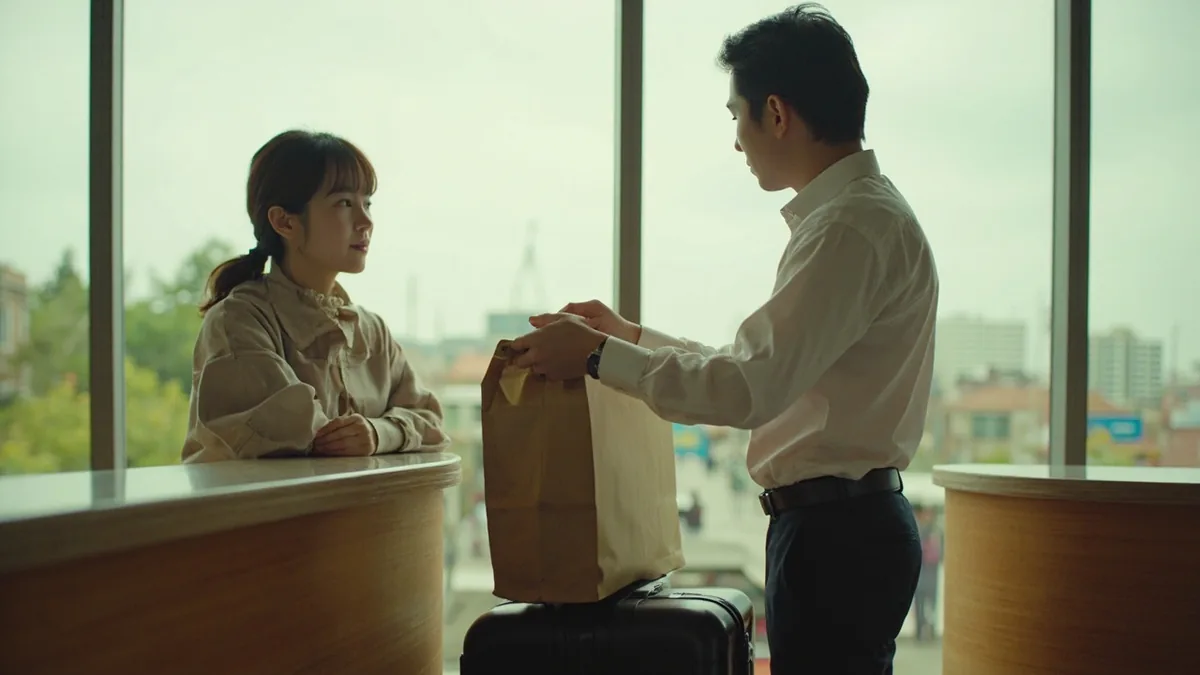 A traveler hands a compact suitcase to a hotel desk attendant, illustrating “please store my luggage” in Chinese: 帮我寄存行李 (bāng wǒ jìcún xíngli)