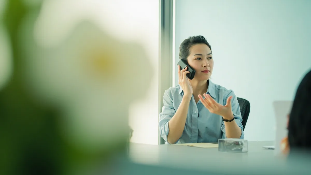 A helpful office worker answers a phone call with a question, illustrating “who would you like to speak to” in Chinese: 请问您找哪位？ (qǐngwèn nín zhǎo nǎ wèi?)
